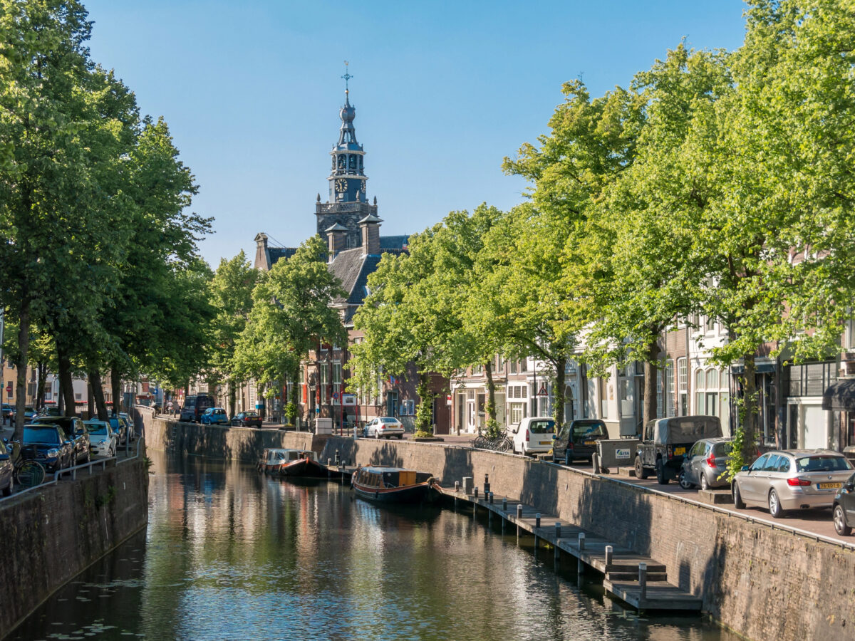 Een Nederlandse gracht in Gouda omzoomd door bomen en gebouwen. In het water weerspiegelen boten en de omgeving, terwijl auto's langs de kade geparkeerd staan.